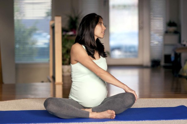 Ethnic mother stretching while sitting on yoga mat at home